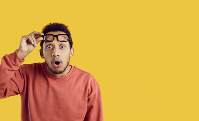 Fototapeta na wymiar Portrait of funny surprised young man looking at you with shocked expression on yellow background. African-American guy in surprise raises his glasses while standing near copy space. Banner.
