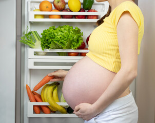 Pregnant woman takes vegetables from the fridge, hungry woman takes carrot from the fridge full of...