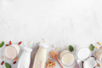 Obraz na płótnie Canvas Vegan, plant based, non dairy milk bottom border. Variety of types in milk bottles and glasses with scattered ingredients. Top view over a white marble background with copy space.