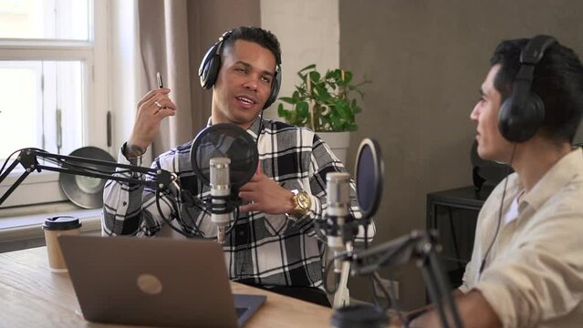 Two businessmen discussing business and recording podcast at table in modern studio spbd. Young Latin men talk and look with smiles, gesticulate with hands and sit at desk in front of microphones in