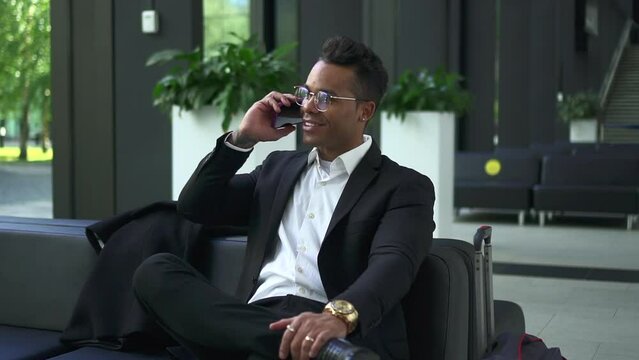 Young businessman having mobile call and waiting for flight while sitting in terminal spbd. Closeup view of African American man talks on phone and looks with happy smile, sits at lounge area, luggage
