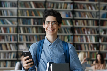Portrait of happy smart Jewish male student in eyeglasses with textbooks and smartphone in hands...