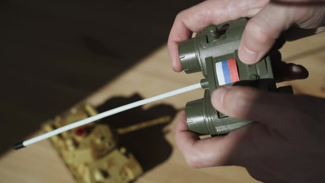 Male hands hold control panel of toy radio-controlled tank with the Russian flag. Blurred war machine toy rides across wooden table. Human fingers move control levers.
