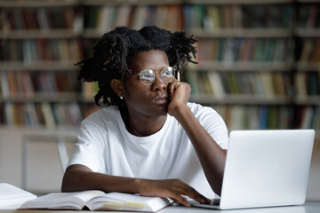 Unhappy young African American hipster guy in eyeglasses looking in distance, feeling bored preparing for examination or doing high school research project on computer, sitting at table in library.
