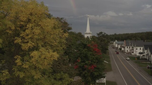 Ascending aerial above Fall colored trees revealing church Monson Maine with rainbow 