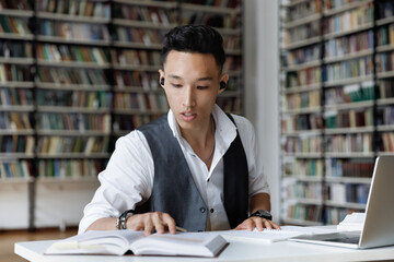 Concentrated millennial male Korean student in earbuds listening favorite lounge music or educational lecture while reading book, preparing for exam or doing research alone in college library.