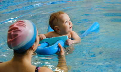 Early age swimming in pool. Baby boy trained to swim in water. Happy child with trainer woman in indoor swimming pool playing and having fun. Healthy and sport family with infant, active parent