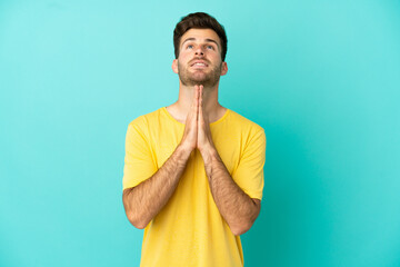 Young caucasian handsome man isolated on blue background keeps palm together. Person asks for...