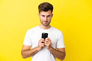 Young caucasian handsome man isolated on yellow background looking at the camera and smiling while using the mobile