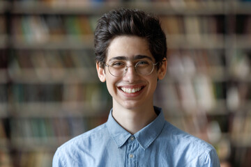 Head shot portrait of happy handsome Jewish male student in eyeglasses posing at blurred library...