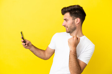 Young caucasian handsome man isolated on yellow background using mobile phone and doing victory gesture