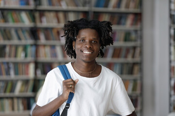 Head shot portrait of smiling 20s African American guy with backpack posing near bookshelves in modern library. Profile photo of happy smart millennial multiethnic male student looking at camera.