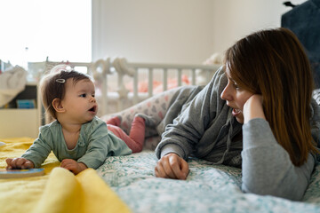 Fototapeta premium Front view on happy small caucasian baby lying on the bed playing with book toy by her mother at home in day copy space childhood growing up family concept