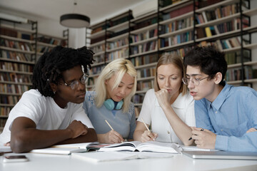 Concentrated group of multiethnic young students reading textbook, writing notes preparing for seminar or exams together, improving knowledge sitting at table in modern library or classroom.