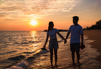Silhouette of happy Asian couple hands holding and walk together on the beach while golden sunset time evening.