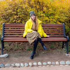 Smiling woman in a yellow jacket sits on a park bench against the backdrop of autumn bushes