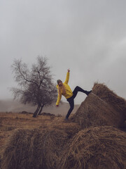 A woman poses with her leg on a haystack and her fingers pointing in different directions in an autumn field against a haze of fog