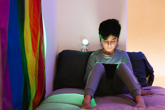 Ethnic boy watching tablet on bed near rainbow flag
