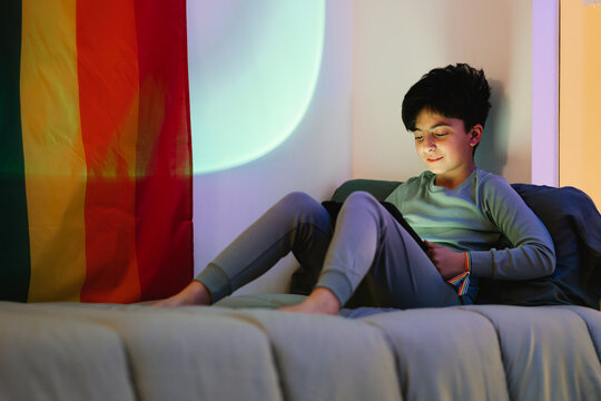 Ethnic boy watching tablet on bed near rainbow flag