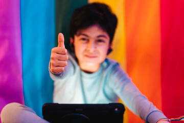 Ethnic boy in earbuds using tablet and showing thumb up