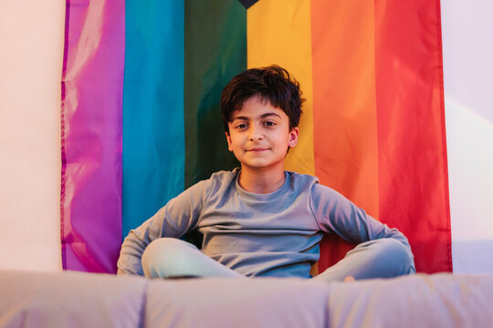 Ethnic boy sitting on bed near rainbow flag