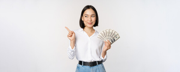Smiling young modern asian woman, pointing at banner advertisement, holding cash money dollars,...