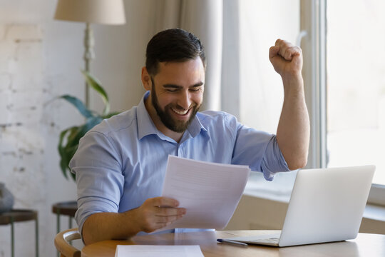 Happy young man sit at desk celebrate great fantastic news in paper formal legal documents read notice raise hand clenched fist looks overjoyed. Success, career advance, business achievements concept
