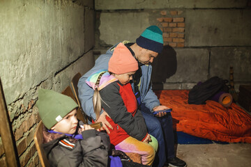 Ukrainian father with two kids sits in a bomb shelter, waits for the end of the air attack of Russian invaders and reads the latest braking news with smartphone. Terrorism and war, current history - obrazy, fototapety, plakaty
