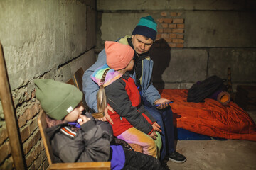 Terrorism and war, current history. Ukrainian father with two kids sits in a bomb shelter, waits for the end of the air attack of Russian invaders and reads the latest braking news with smartphone