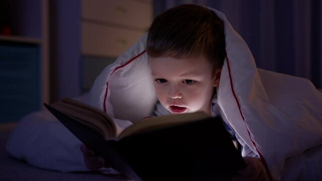 Small blonde caucasian boy of 5 years old lies on floor covered with white blanket in dark bedroom, reading book with fairy tales and light from night lamp. Small boy studying book in evening at home.