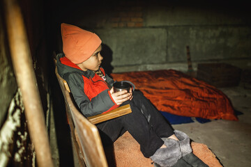 Little Ukrainian girl sits in a bomb shelter, drinks tea from a thermos and waits for the end of...