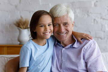 Portrait of grey-haired grandfather and little school age grandson hugging seated on cozy couch at...