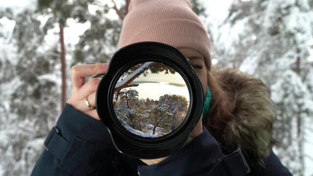 A young woman photographer takes a picture in a winter forest, snow-covered pines and sunset are reflected in the lens as in a mirror.