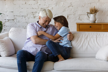 Older 60s grandfather have fun play with little cute grandson, kid and mature granddad tickling each other look happy, laughing sit on sofa at home. Multi generational family playtime, leisure concept
