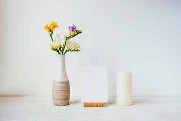 Blank paper, candle and bouquet of white, yellow and blue freesia flowers in vase on white background. Home decor, mock up