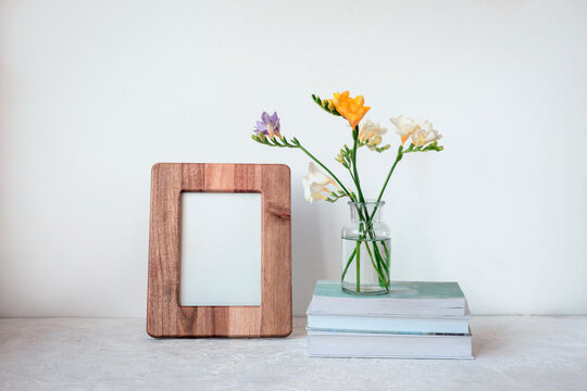 Blank picture frame and flowers in vase standing on stack of books on white background. Front view, mock up