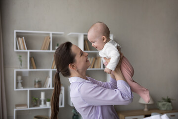 Cheerful happy new mom lifting adorable baby, holding infant in arms, looking at little laughing son, daughter, smiling, talking to child. Young mother cuddling new born kid at home