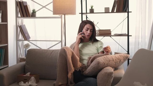 Unexpected disease. Sick woman. Distance support. Upset lady covering plaid sitting sofa telling about high temperature on smartphone in light room interior.