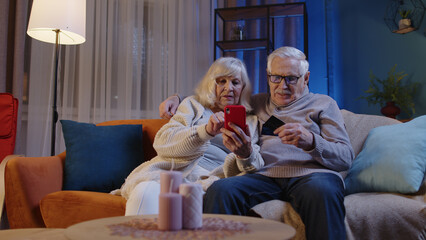 Happy smiling senior family couple with smartphone on sofa at home, making online shopping with...