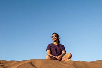 Young man with long black hair wearing glasses at the beach sitting on the sand with a blue sky at background