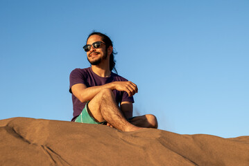 Young man with long black hair wearing glasses at the beach sitting on the sand with a blue sky at background