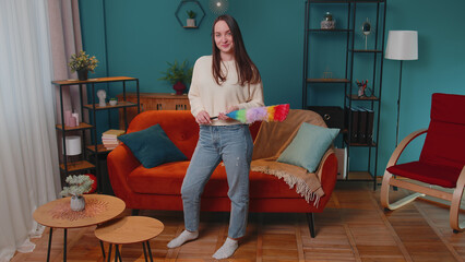 Happy one young woman dusting cleaning furniture caring for hygiene using colorful duster in living room. Girl housekeeper wiping dust from sofa and table at home. Housework and domestic life concept