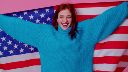 Trendy cheerful positive teen girl waving and wrapping in American USA flag, celebrating, human rights and freedoms. Independence day. Young pretty adult woman. Indoor studio shot on pink background