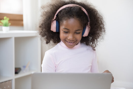 Happy Adorable Little African American Girl In Wireless Headphones Watching Funny Educational Online Lecture On Computer, Enjoying Learning Foreign Language Or Studying On Online Courses Alone At Home