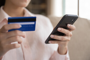 Close up happy young female client holding bank card and smartphone in hands, making payments using mobile e-banking application, shopping online in internet store, purchasing goods or services.