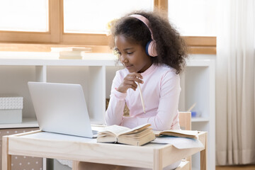 Happy concentrated adorable small African American child girl in headphones looking in laptop...
