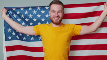 Portrait of bearded young man in t-shirt waving, wrapping in American USA flag, celebrating, human rights, freedoms. Independence day. Adult guy indoors studio shot isolated alone on gray background