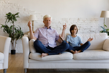 Grandfather and little grandson with eyes closed do meditation practice sit on sofa in living room,...