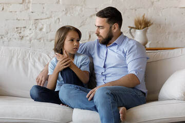 Serious father listen to his pre-teen little son talking seated on sofa at home, speaking spend...