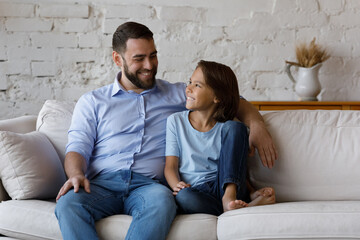 Young dad his lovely smiling little son sit on sofa, talking, hugging enjoy carefree weekend together at modern home. Understanding communication of friendly wellbeing family, Happy Father Day concept